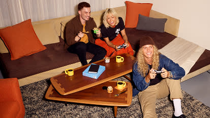 Three friends relaxing on a beige sectional sofa with orange pillows, drinking from yellow mugs around a wooden coffee table