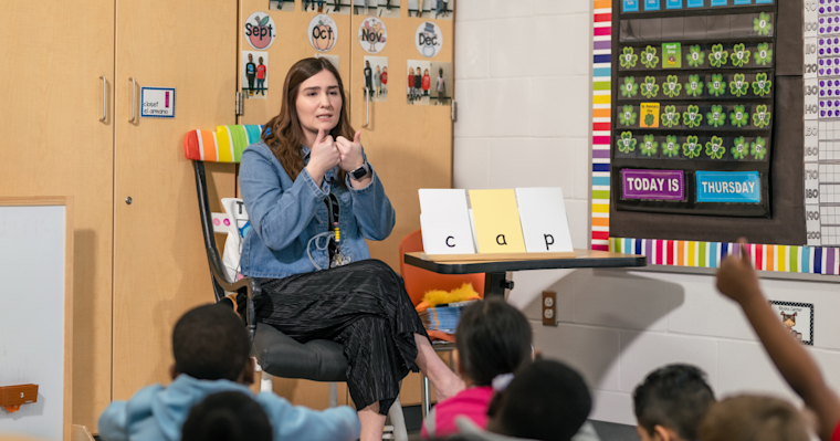 A teacher sits at the front of a classroom leading a phonics lesson with young students. She gestures with her hands while facing the class. On a small table beside her are letter cards spelling “cap.” 