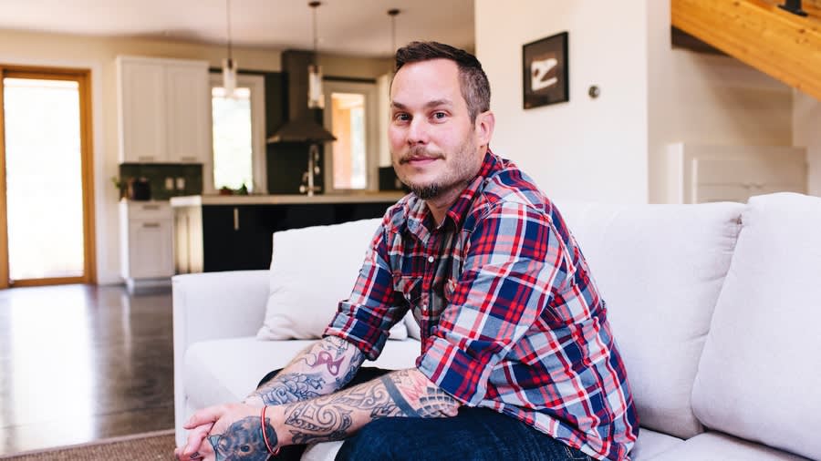 Person in plaid shirt with tattoos sitting on white couch in modern open-concept living room with kitchen visible in background