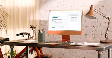 Modern desk workspace with copper desk lamp, keyboard, and email campaign interface displayed on screen against white brick wall