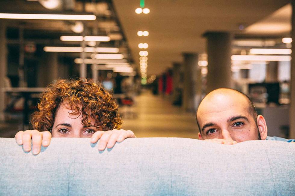 Two office workers playfully peek over a cubicle wall in an open office space with fluorescent lighting overhead