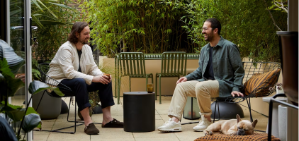 Two people sitting outdoors in patio chairs having an animated conversation, with a dog resting nearby on a sunny day