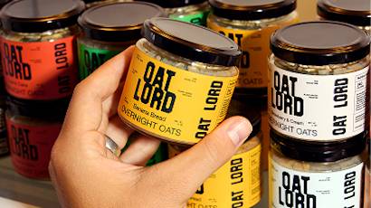 Glass jars with black lids lined up on shelf, showing yellow and black labels for oat-based products