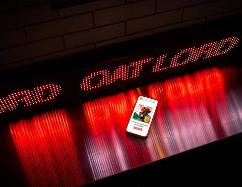 Smartphone displaying Oat Lord app interface on reflective surface with red LED text display and dramatic red lighting effects above