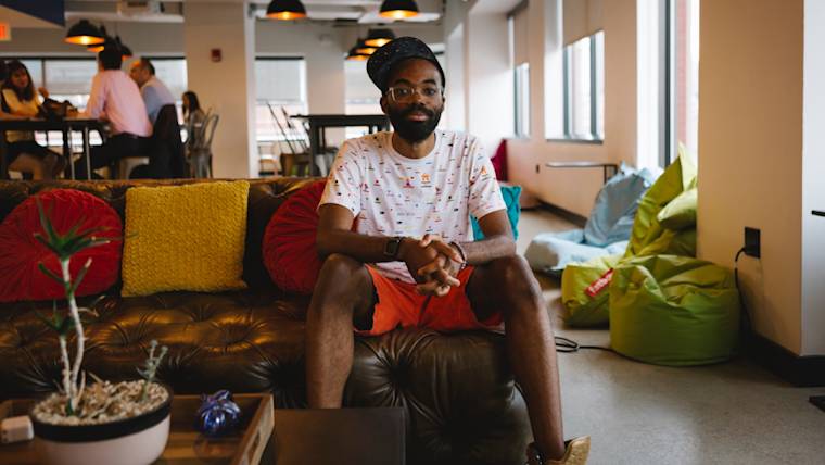 Web Designer Paul Anthony Webb relaxes on a leather couch in a modern office lounge with colorful pillows and pendant lighting overhead
