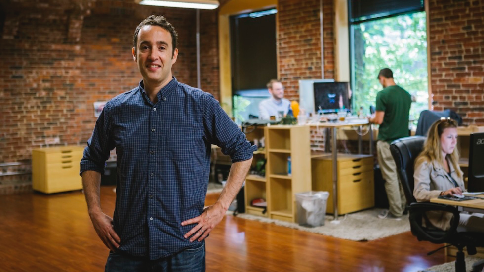 Professional in blue checkered shirt standing in modern office with exposed brick walls and coworkers working in background