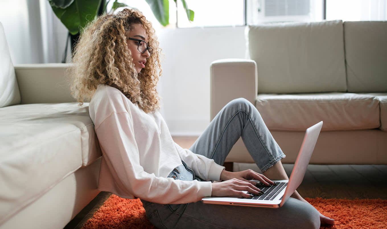 A young woman with curly hair sitting on the floor against a couch typing on her computer. She is sitting on an orange rug and there's a plant behind her.