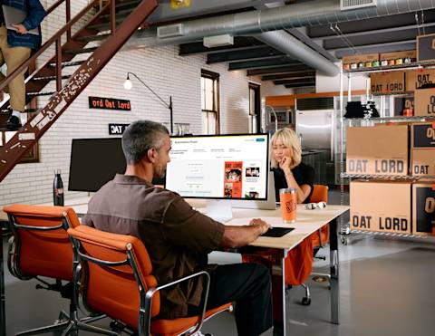 Two people collaborating at a modern office workspace with Oat Lord branding visible on walls and computer screens