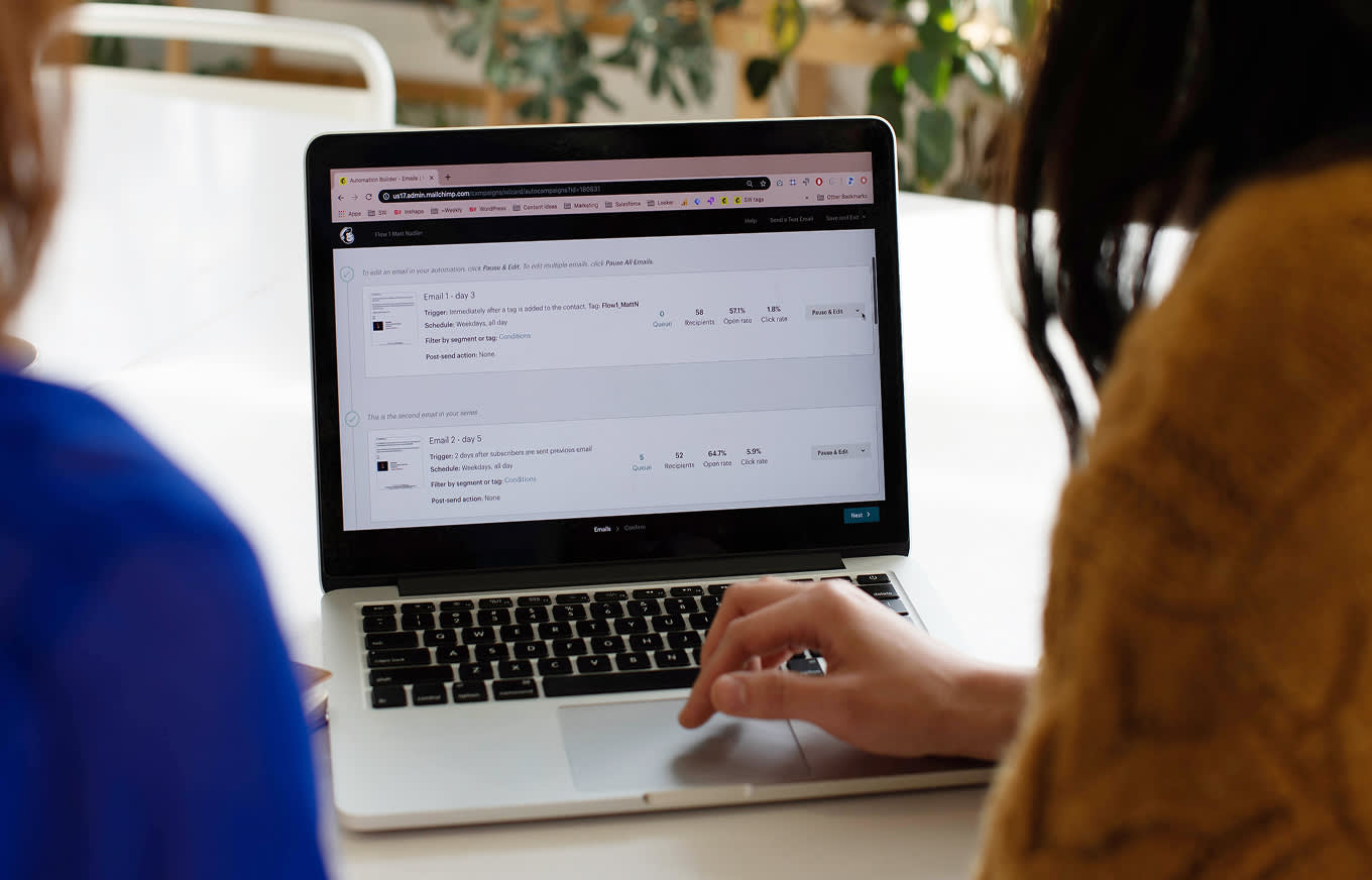A person's hand is using a laptop showing the Mailchimp app on the email automation screen with two emails and analytics for the automated emails. There's another person sitting next to them. There are plants behind the table.