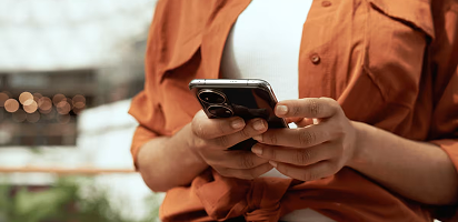 Person in orange shirt using smartphone, close-up view of hands texting on mobile device outdoors
