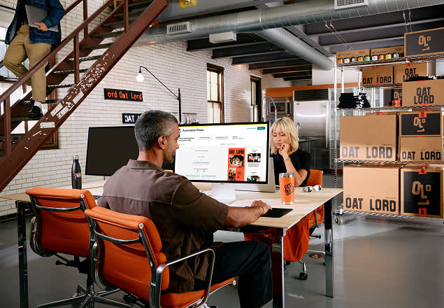 Two people working in a modern industrial office space with white brick walls, orange chairs, and Oat Lord branding displayed throughout