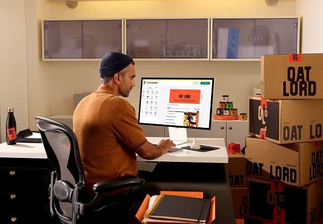 Designer in brown shirt working at desk with computer monitors, surrounded by branded Oat Lord packaging boxes