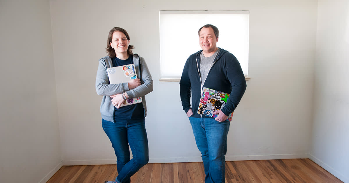 Two software developers standing in empty room with laptops decorated with stickers, wearing casual hoodies and jeans near window