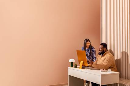 Two colleagues smiling and collaborating at a desk with a computer monitor in a warm, modern workspace.