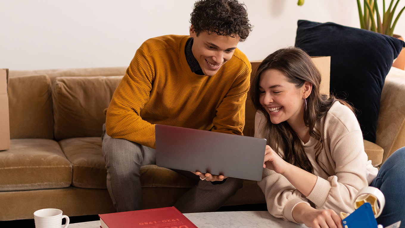 A young man and a young woman sitting on and beside a couch looking at a laptop screen and smiling. They are in front of a coffee table with a book and a coffee mug.