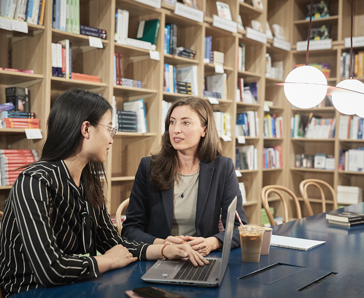 Two professionals having a discussion at a table with laptops and coffee in a modern library or bookstore setting with wooden shelving
