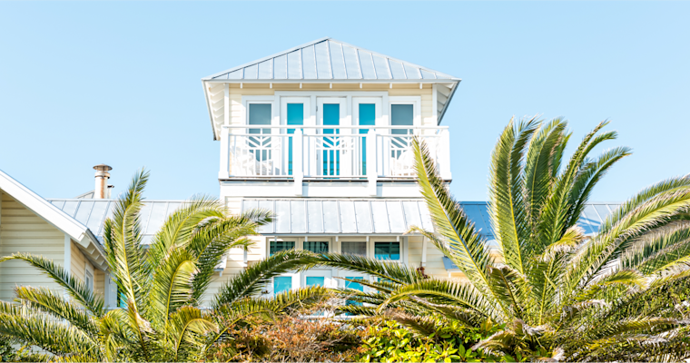 White coastal beach house with turquoise windows and metal roof, framed by lush palm trees against bright blue sky