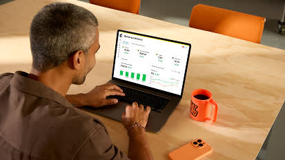 Person working at wooden desk with laptop, orange coffee mug, and black notebook in modern office setting