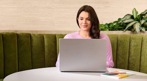 Woman in pink shirt, sitting on a green couch, working on a laptop