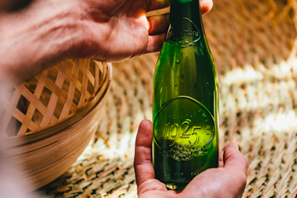 Hands holding a green glass beer bottle against a woven bamboo basket background, creating a warm, rustic atmosphere