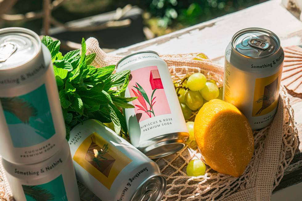 Premium soda cans from "Something & Nothing" brand arranged with fresh mint, lemon, and grapes on a woven cloth in sunlight