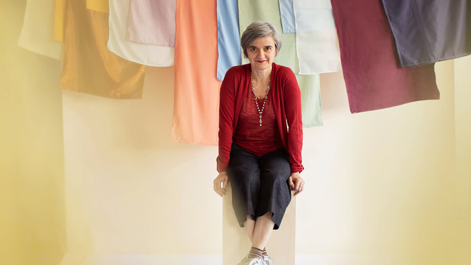 A photograph of a woman sitting on a stool in front of colorful linens.