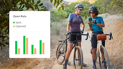 Two cyclists in athletic gear pause on a dirt trail to check their phones, with bikes and a scenic hillside backdrop
