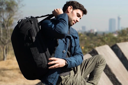 Person wearing navy jacket adjusts large black backpack outdoors with city skyline visible in background