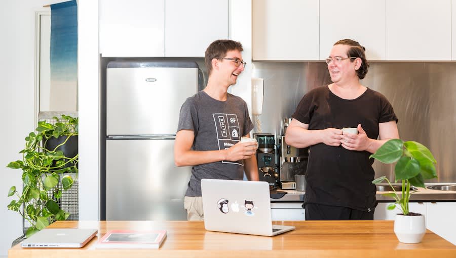 Two colleagues share a laugh while having coffee in a modern kitchen with stainless appliances, laptops, and potted plants