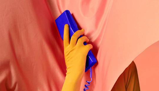 Hand wearing yellow rubber glove holding blue retro telephone handset against coral pink background