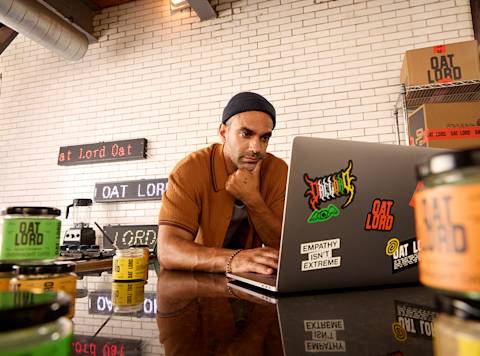 Person working on laptop in warehouse setting surrounded by Oat Lord branded products and stickers against brick wall background
