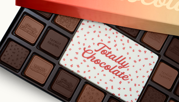 Open gift box of assorted chocolates from Totally Chocolate, featuring individually molded chocolate squares surrounding a large white chocolate centerpiece printed with the brand name. The chocolates are arranged in a black tray inside a red and gold box.