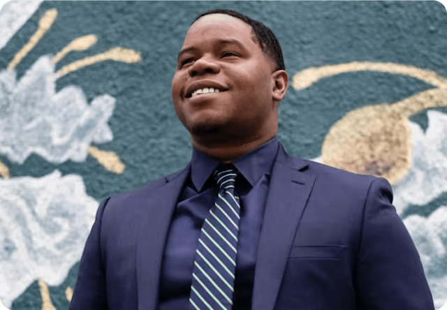 Immagine di Dundalan Sain, Direttore del Supporto Franchising, vestito in abito e in piedi davanti a un murale di fiori colorati dipinto su una parete