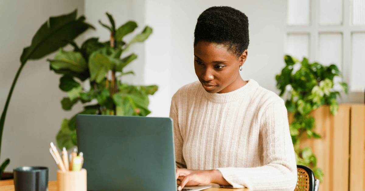 A woman wearing a white sweater works at a table, typing on a laptop keyboard with indoor plants in the background, and a drink within reach.