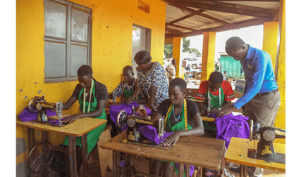 Students in green aprons learn sewing at wooden tables with vintage machines in a yellow-walled classroom, working with purple fabric