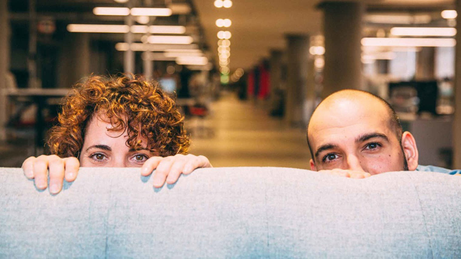 Two office workers playfully peek over a cubicle wall, one with curly hair and one bald, in a warmly lit workspace