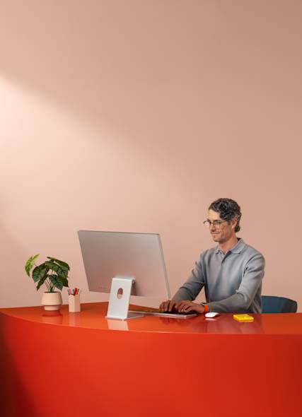 A man wearing glasses working at a desktop computer on a bold orange desk in a modern, minimalist workspace.