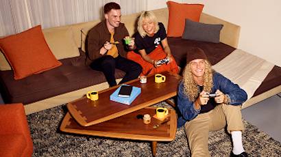 People relaxing on a gray couch with orange pillows, enjoying drinks and snacks while socializing in a casual home setting