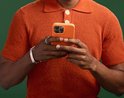 Hands holding an orange smartphone, wearing silver bracelet, against orange polo shirt and green background