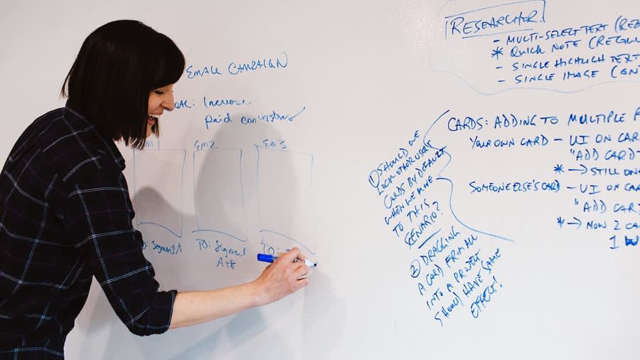 Person in black plaid shirt writing on whiteboard with blue marker, surrounded by project notes and diagrams