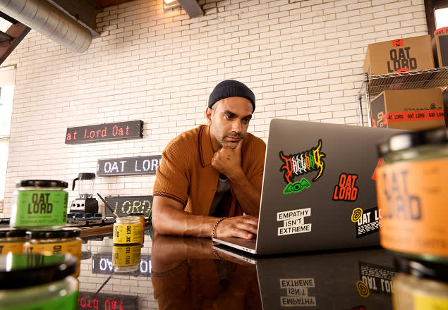 Person in brown shirt working on laptop surrounded by Oat Lord branded products in office setting with white brick wall background