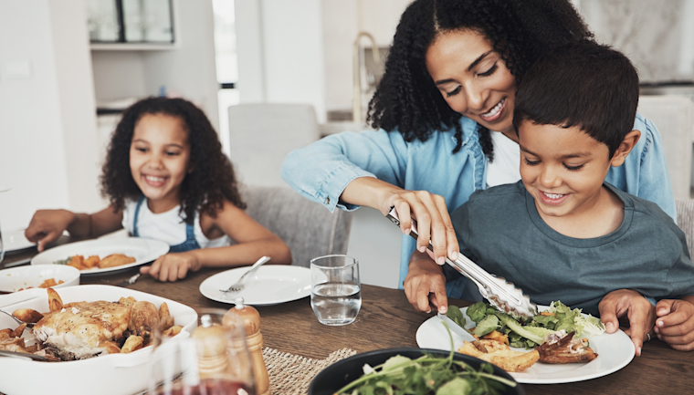 Family gathered around dining table sharing a meal together, smiling while reaching for food on plates with salad and bread