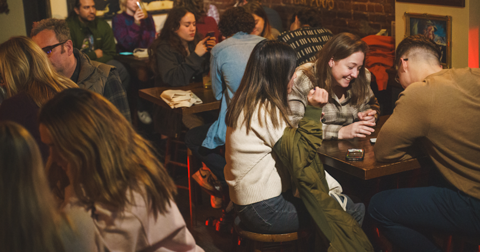 A crowded bar during a trivia night, with groups of adults seated at small wooden tables, talking, laughing, and looking at phones and notes.