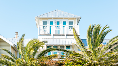 Row of white coastal homes with metal roofs seen through tropical palm fronds against bright blue sky