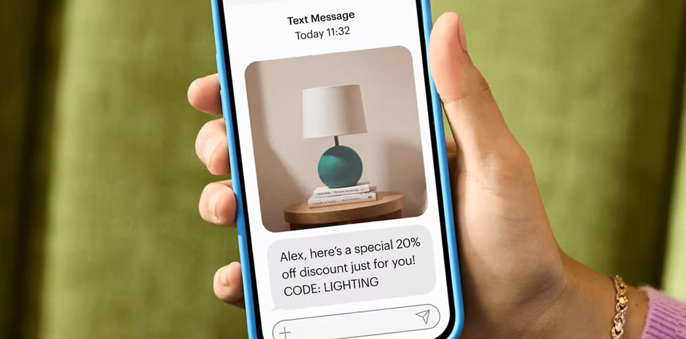 A person's hand holding up a phone with a text message that shows an image of a teal lamp on a small table, on top of a stack of books. Beneath the image, the text reads "Alex, here's a special 20% off discount just for you! CODE: LIGHTING"