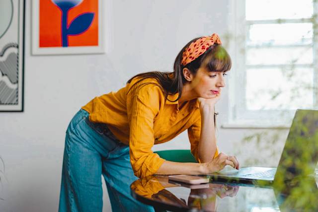 Person in yellow blouse and denim overalls working at desk with laptop, wearing orange patterned headband in bright modern office space