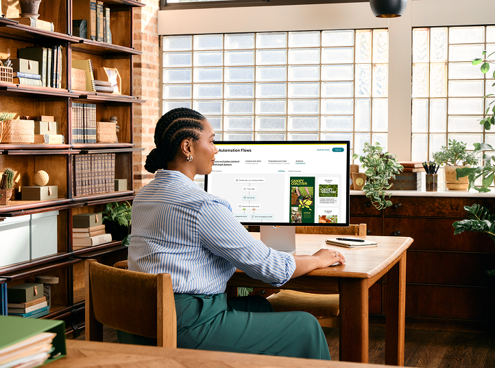 Professional working at wooden desk in library-like setting with bookshelves and plants, viewing computer screen in natural light