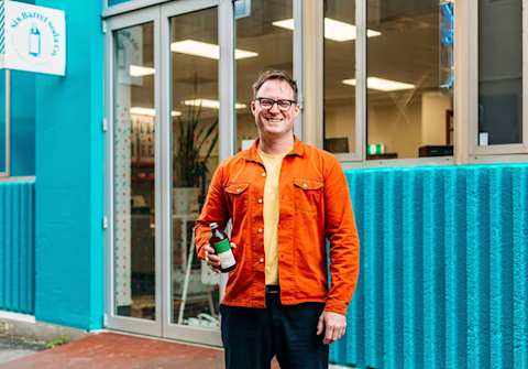 Customer in bright orange jacket and yellow shirt holding a bottle outside storefront with vibrant turquoise walls