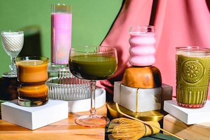 Colorful drinks in various glasses arranged on white pedestals against pink and green backdrop, with decorative props and gold whisk