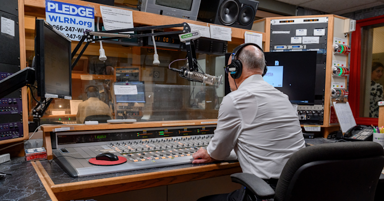 A radio host wearing headphones sits at a large audio mixing console inside a broadcast studio. He faces a microphone and computer monitors, with equipment racks and speakers surrounding the workspace.